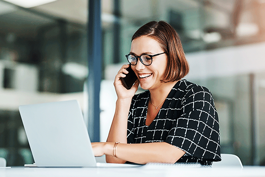 Nainen puhuu kännykkään ja käyttää kannettavaa tietokonetta toimistossa